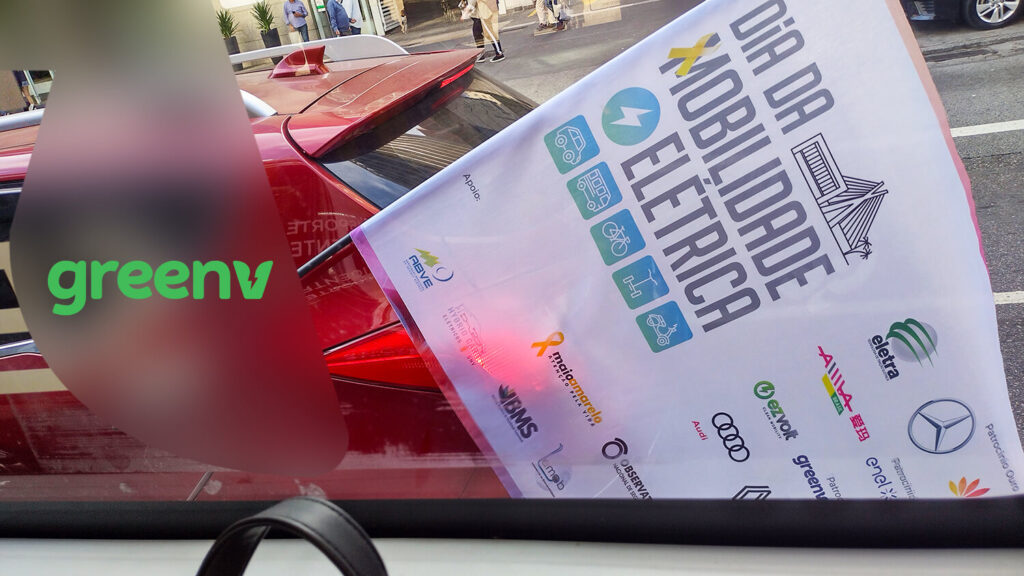 Temos a foto de uma bandeira com os dizeres “Dia da Mobilidade Elétrica” e os ícones dos patrocinadores, sua haste está encaixada na janela traseira de um carro vermelho.