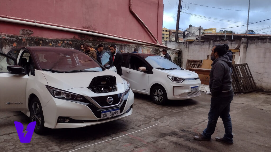 Temos um modelo branco do Nissan Leaf ao lado de um JAC E-JS1, também na coloração branca. Ao redor dos veículos elétricos temos alguns participantes da palestra sobre Introdução a Eletromobilidade.