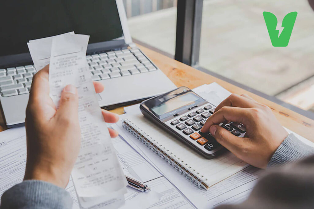 Fotografia colorida mostra uma pessoa conferindo valores de uma nota fiscal na calculadora.
